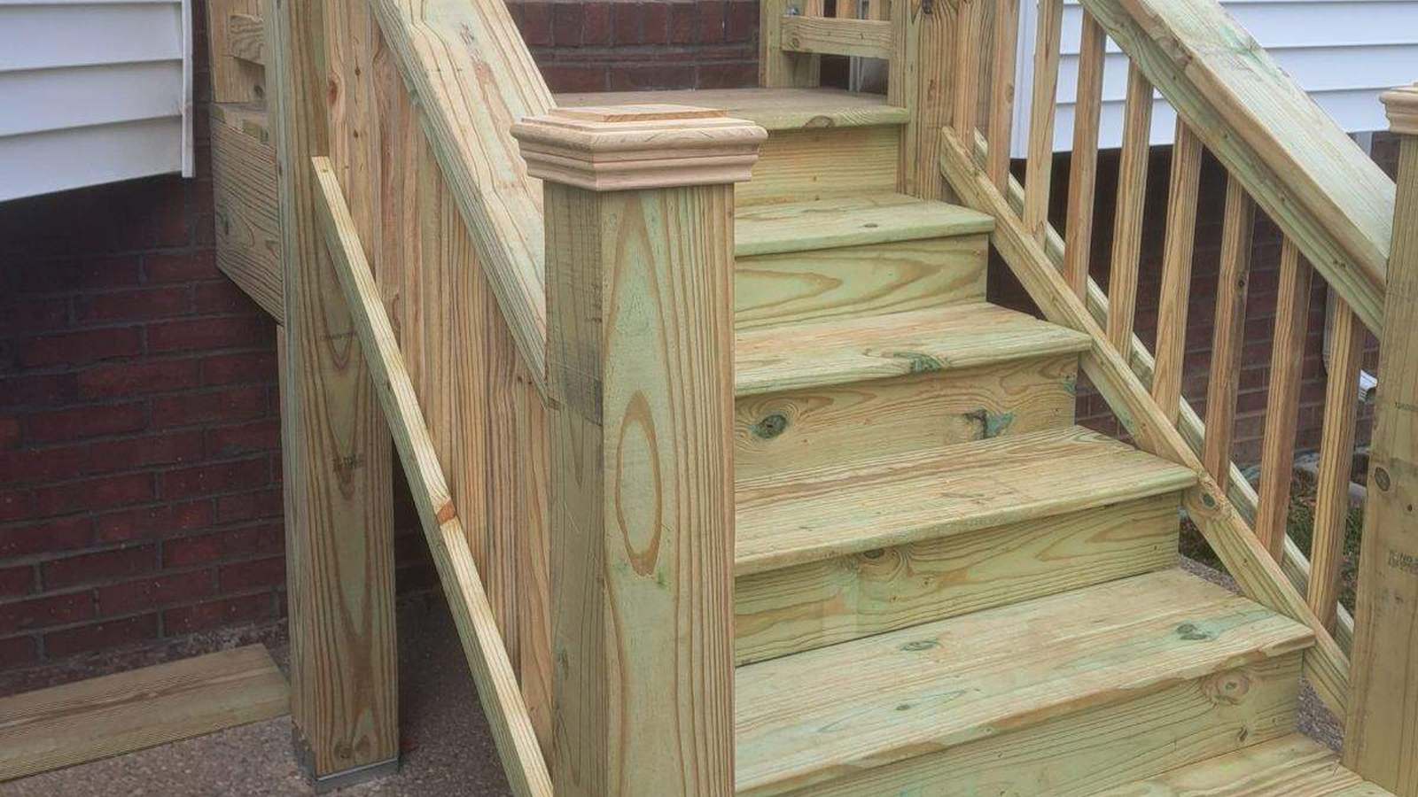 A newly built pressure-treated deck stair and railing at the back door of a brick and vinyl-sided house, with fresh-cut treads and turned post caps.
