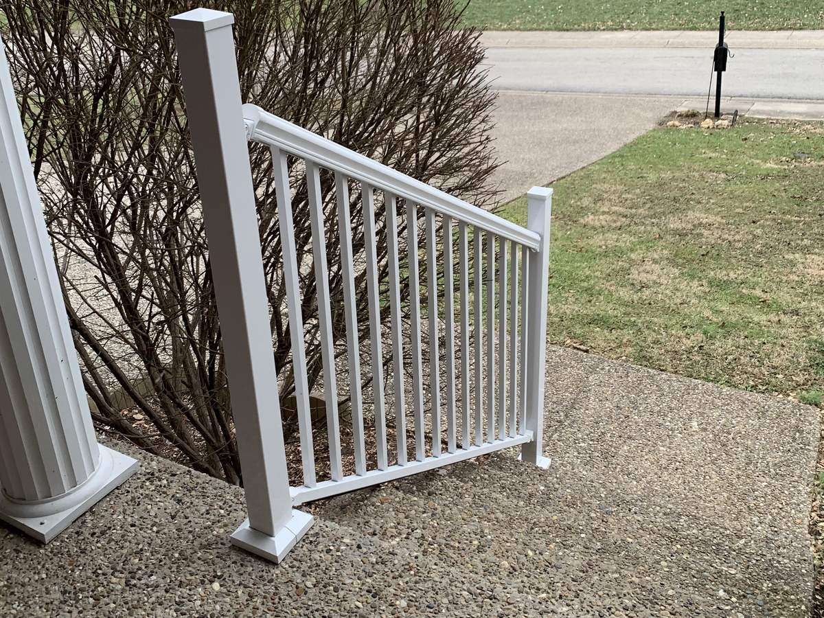 A newly installed white railing with square posts on a concrete front porch step, beside a white porch column, with a lawn and street behind.